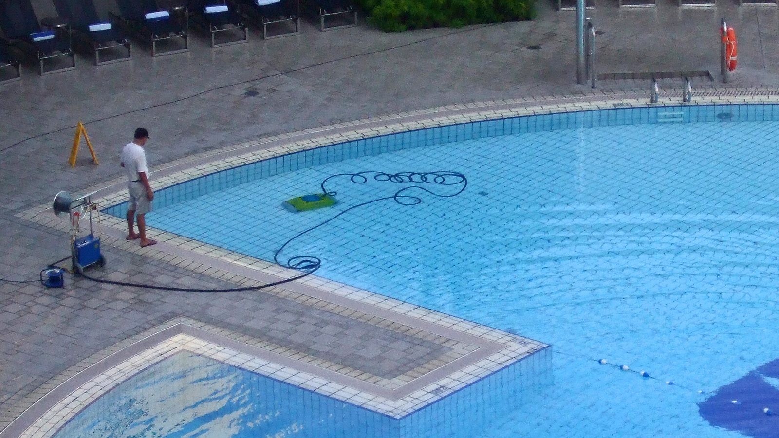 A man on a pool deck watching a corded robot clean the pool floor